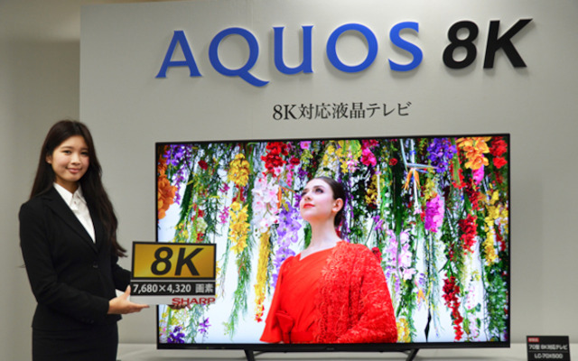 a lady stands next to a large television holding an 8K sign